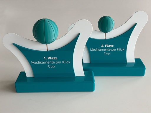 Zwei Awards für den Medikamente Per Click Cup aus türkisem Acrylglas mit Lasergravur. Die Gravur wurde mit weißem Lack ausgelegt. Die türkisen Seitenflächen wurden beidseitig auf eine weiße Acrylglas-Trägerplatte geklebt. Die Köpfe der stilisierten Figuren sind aus 3D-gedrucktem PETG Material und sehen aus wie Basketbälle. Der Kopf des Awards für den zweiten Platz ist kleiner als der des ersten Platzes.