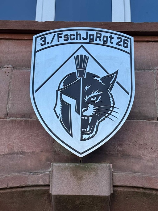 Ein 6 mm Edelstahlschild in Wappenform, auf das ein 3 mm schwarz-pulverbeschichteter Edelstahlschriftzug und ein Logo einer Bundeswehrdivision geschraubt wurde. Das Wappen wurde mit 4 mm abgekanteten Edelstahlhaltern an einer Kalksandsteinfasse befestigt.