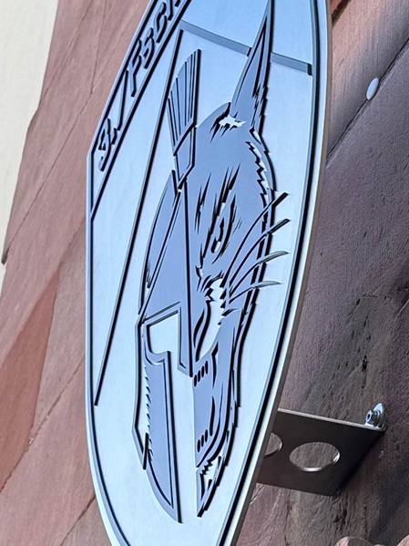 Ein 6 mm Edelstahlschild in Wappenform, auf das ein 3 mm schwarz-pulverbeschichteter Edelstahlschriftzug und ein Logo einer Bundeswehrdivision geschraubt wurde. Das Wappen wurde mit 4 mm abgekanteten Edelstahlhaltern an einer Kalksandsteinfasse befestigt.