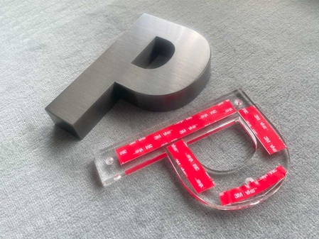 Profilbuchstabe mit Brush Black Finish, daneben derselbe Buchstabe aus 10 mm klarem Acrylglas mit Senklochbohrungen und Doppelseitigem Klebeband für eine "schwebende" Befestigung