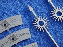 Foto zeigt einzelne Edelstahl-Bestandteile einer Sonnenuhr die laserbeschriftet sind. Die Schilder zeigen Texte wie "Wahrer Mittag" oder Wolkensymbole etc. In die Zeiger ist ein Sonnensymbol integriert. Alle Teile liegen auf einem blauen Hintergrund.