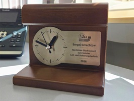Foto zeigt eine Tischuhr mit einem Holzkörper. Auf der Vorderseite ist ein Edelstahlschild mit schwarz-matter Laserbeschriftung aufgeschraubt. Die Laserbeschriftung ist sowohl das Ziffernblatt als auch die Widmung für eine bestandene Meisterprüfung.
