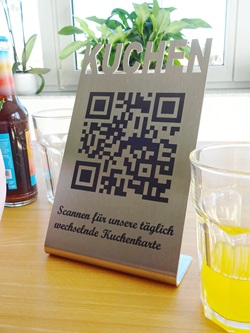 Foto zeigt einen aus Edelstahl abgekanteten Tischaufsteller mit Laserbeschriftung. Oben am Edelstahlblech ist das Wort "KUCHEN" Teil der Außengeometrie. Darunter steht ein QR-Code. Darunter steht "Scannen für unsere täglich wechselnde Kuchenkarte". Das Schild steht auf einem Holztisch mit mehreren Gläsern und einer Limonadenflasche. Im Hintergrund sieht man zwei Fenster und mehrere Pflanzen auf einer Fensterbank.