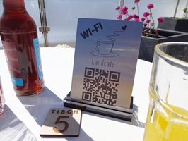 Foto zeigt einen Tischaufsteller aus Edelstahl mit Laserbeschriftung, der in einem Acrylglas-Block steht. Oben auf dem Schild steht WI-FI, eine Kaffeetasse und darunter das Wort Landcafé. Darunter steht ein QR-Code. Das Schild steht draußen auf einem weißen Tisch mit mehreren Gläsern und einer Limonadenflasche. Im Hintergrund sieht man mehrere Pflanzenkübel und einen Sichtschutz aus mattiertem Acrylglas.