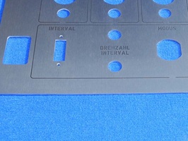 Edelstahlzuschnitt mit mehreren runden und rechteckigen Ausschnitten. Das Schild wurde laserbeschriftet, um an den Ausschnitten die Funktionen der einzubauenden Schalter zu zeigen. Der Zuschnitt liegt auf einem blauen Hintergrund.