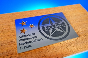 Foto zeigt einen rechteckigen Edelstahlzuschnitt mit anthrazit-farbener Laserbeschriftung. Im linken oberen Bereich sind drei Sterne ausgeschnitten. Darunter steht der Text "Astronomiewettbewerb Niedersachsen 1. Platz". Auf der rechten Hälfte ist ein großer Stern auf dem mittig "Astro Stars" steht. Das Schildchen liegt auf einem Holzblock.