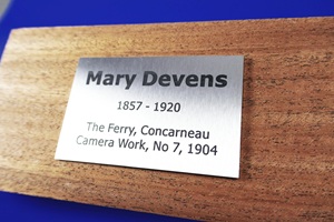 Foto zeigt einen rechteckigen Edelstahlzuschnitt mit anthrazit-farbener Laserbeschriftung. Das Schild ist eine Ehrenplakette für Mary Devens, eine berühmte US-amerikanische Fotografin des 19. Jahrhunderts. Das Schild liegt auf einem Holzblock.
