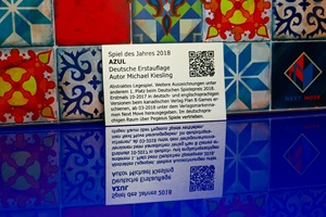 Foto zeigt einen rechteckigen Edelstahlzuschnitt mit anthrazit-farbener Laserbeschriftung. Auf dem Schild wird das Brettspiel "Azul" beworben. Im Hintergrund ist der untere Abschnitt des Brettspielkastens für Azul zu sehen.