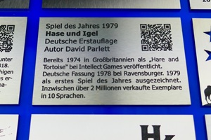 Foto zeigt einen rechteckigen Edelstahlzuschnitt mit anthrazit-farbener Laserbeschriftung. Auf dem Schild wird das Brettspiel "Hase und Igel", Spiel des Jahres 1979, beworben. Um das Schild herum liegen im Raster noch weitere, gleich große Edelstahlschilder mit Laserbeschriftung. Schild liegt auf blauem Untergrund.