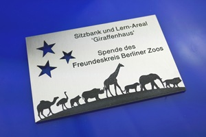 Foto zeigt einen rechteckigen Edelstahlzuschnitt mit anthrazit-farbener Laserbeschriftung. Im linken oberen Bereich sind drei Sterne ausgeschnitten. Rechts daneben steht "Sitzbank und Lernareal Giraffenhaus. Spende des Freundeskreises Berliner Zoo". Unten an der Kante steht eine Karawane aus vielen afrikanischen Tierarten. Schild liegt auf blauem Untergrund.