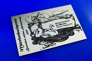 Foto zeigt einen rechteckigen Edelstahlzuschnitt mit anthrazit-farbener Laserbeschriftung. Seitlich steht senkrecht der Text "Märchenbuchpreis 'Besonders schöne Ausstattung' 2019", rechts daneben sind zwei Schwäne abgebildet die in einem Teich schwimmen, im Hintergrund thront ein Schloss auf einer Burg. Neben den Schwänen ist ein Banner auf dem "Michael Köhlmeier, Die Märchen. Hanser Verlag, 2019". Schild liegt auf blauem Untergrund.