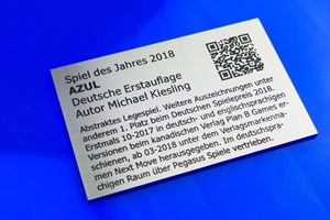 Foto zeigt einen rechteckigen Edelstahlzuschnitt mit anthrazit-farbener Laserbeschriftung. Auf dem Schild wird das Brettspiel "Azul" beworben. Schild liegt auf blauem Untergrund.