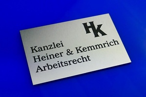 Foto zeigt einen rechteckigen Edelstahlzuschnitt mit anthrazit-farbener Laserbeschriftung. Darauf ist das Logo und der Text für eine Kanzlei für Arbeitsrecht abgebildet. Schild liegt auf blauem Untergrund.