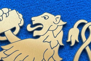 Foto zeigt einen Edelstahl-Laserzuschnitt mit einem heraldisch-anmutenden Löwen, der einen Bierkrug hält. Der Zuschnitt wurde TiN-Gold beschichtet und anschließend laserbeschriftet. Der Löwe liegt auf blauem Untergrund.